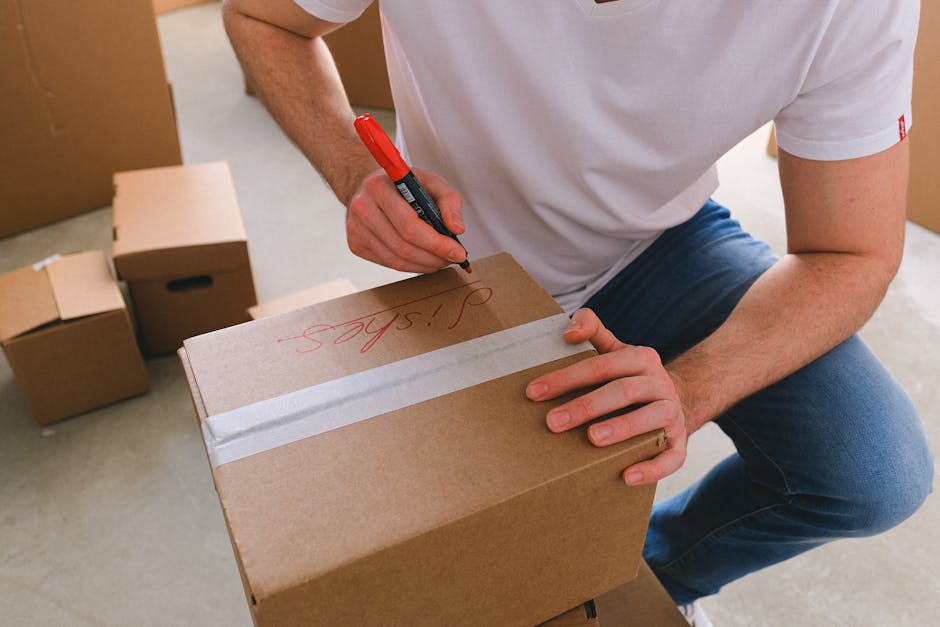 Close-up of a person in a white t-shirt and jeans, kneeling on the floor inside a property during a home relocation process, marking a cardboard moving box with a red marker. The individual is writing the word 'Spares' on the box, which is secured with white packing tape across the top. Surrounding the person are several other cardboard boxes of various sizes, some stacked or placed on the floor, indicating packing and moving preparations. The scene shows a well-lit indoor environment with a neutral floor surface and open doorway in the background, suggesting the area near an exit or doorway used for loading furniture and belongings. The presence of packing materials and the action of labeling boxes align with professional furniture transport and packing services provided by Man with Van Shacklewell during house removals.