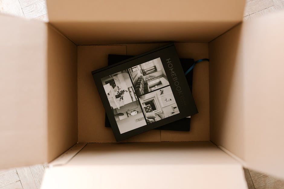 A young man with light brown hair and a beard, dressed in a blue hoodie and dark work trousers, is sitting on the edge of a moving van with open cardboard boxes around him. He is holding and reading a