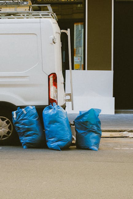 The rear side of a white moving van parked on a street outside a building with a large window, showing three blue plastic rubbish bags filled with bulky waste placed on the pavement next to the vehicle. The van has an open rear door and a roof rack, indicating it is used for home relocation and furniture transport. In the background, a white panel or large piece of foam is positioned against the building’s exterior wall near the door. The scene depicts a packing and loading process typical of house removals, with the waste bags prepared for disposal or collection, supporting the overall theme of professional moving services provided by Man with Van Shacklewell, as referenced on the webpage about bulky waste and sofa disposal in Shacklewell.