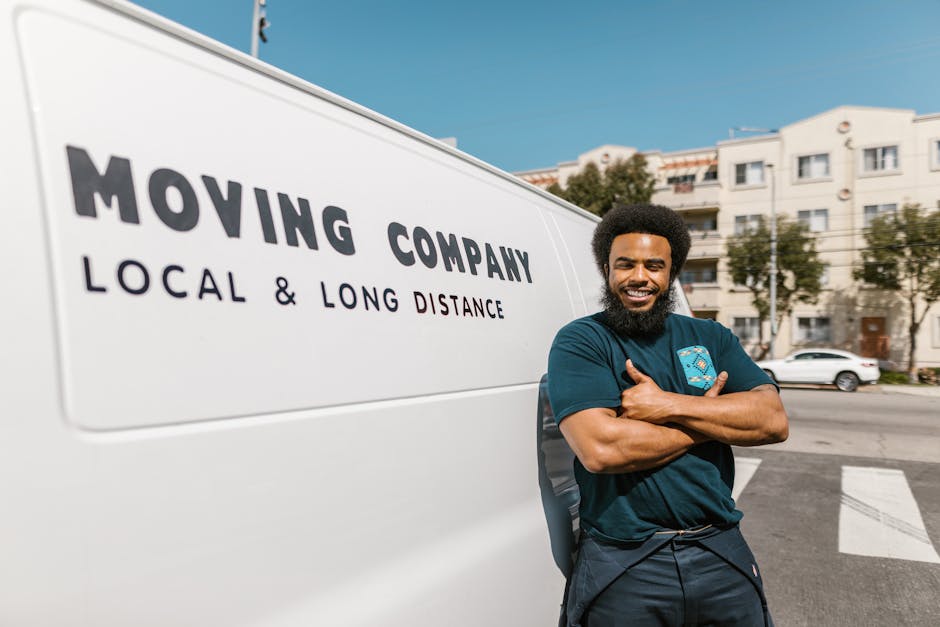 A smiling man with an Afro hairstyle and a full beard leaning against a white moving van on a city street. The van displays the text 'MOVING COMPANY' and 'LOCAL & LONG DISTANCE' in bold black letters. The man is dressed in a navy blue t-shirt with a logo on the chest, and his arms are crossed. Behind him, residential buildings with multiple stories and balconies, as well as parked cars, are visible under a clear blue sky. The scene captures the loading process and professional furniture transport services offered by Man with Van Shacklewell, located near Shacklewell in Hackney, pertinent to house removals and relocation logistics.