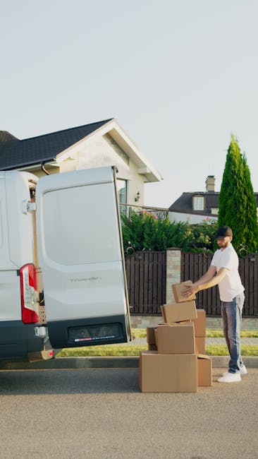 A man wearing a white t-shirt and jeans is standing outdoors on a residential street during daylight, preparing cardboard boxes for loading onto a white moving van. He is holding a cardboard box, with three additional boxes stacked beside him on the pavement. The open rear doors of the van reveal a partially visible interior for furniture and packing materials. The setting includes a wooden fence, a well-maintained garden with green shrubs and a tall, narrow conifer tree, and a house with a gabled roof in the background. The scene captures the packing and loading process involved in home relocation services, with clear emphasis on furniture transport and careful handling of household items by Man with Van Shacklewell, supporting efficient removals and moving logistics in the area of Hackney.