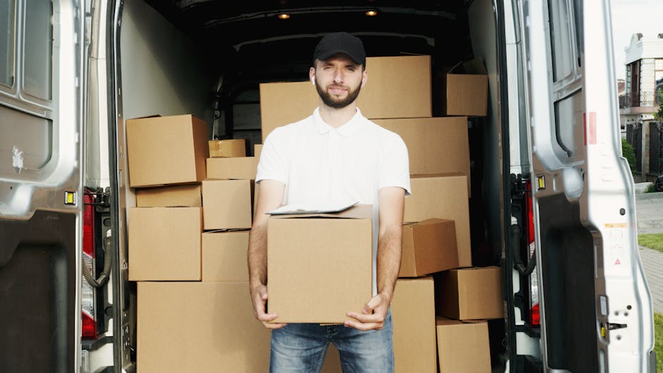 A smiling man with an Afro hairstyle and a full beard leaning against a white moving van on a city street. The van displays the text 'MOVING COMPANY' and 'LOCAL & LONG DISTANCE' in bold black letters. The man is dressed in a navy blue t-shirt with a logo on the chest, and his arms are crossed. Behind him, residential buildings with multiple stories and balconies, as well as parked cars, are visible under a clear blue sky. The scene captures the loading process and professional furniture transport services offered by Man with Van Shacklewell, located near Shacklewell in Hackney, pertinent to house removals and relocation logistics.