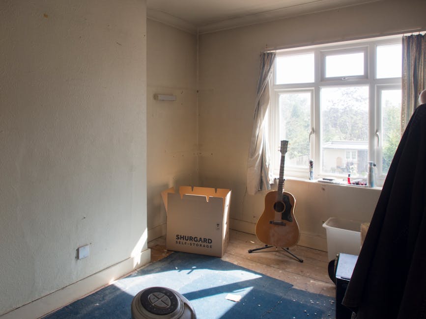 A room with beige walls and a large window allowing natural light to illuminate the space, showing a partially packed home with a cardboard box labeled 'Shurgard Self-Storage' placed on the floor near the wall. A wooden acoustic guitar leans against the window sill, and a white storage bin is positioned to the right of the guitar. The floor appears to be wooden, with a small section of blue carpet visible, and a vacuum cleaner is seen in the foreground on the lower left. Curtains are drawn to the sides of the window, revealing a view of trees and neighboring houses outside. The scene suggests an ongoing house relocation process, with items being prepared for moving or packing, consistent with furniture transport and packing activities that are typical of house removals by Man with Van Shacklewell.