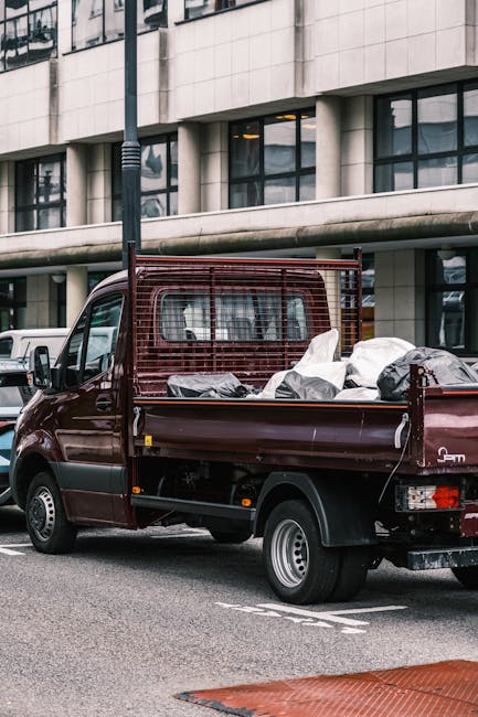 A small, dark red flatbed van parked on a city street in front of a modern building with large glass windows and white stone panels. The van's cargo area is loaded with various household items wrapped in plastic and fabric, including cardboard boxes and furniture. The items are stacked and secured within the open rear section, which features a metal grid partition behind the cab. Nearby, a white vehicle is visible in the background. The scene captures the loading process involved in home relocation or furniture transport, with the van positioned close to the curb on an asphalt road, under overcast lighting. This setup is typical of professional removal services such as those provided by Man with Van Shacklewell, specialising in house removals within the N16 area.