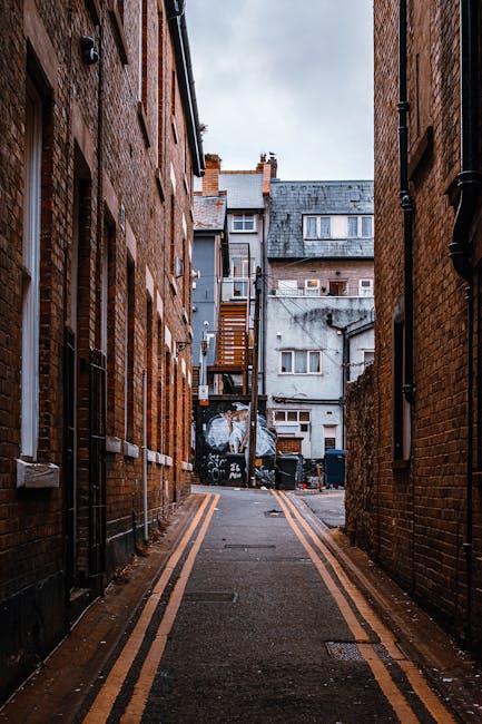 A narrow alleyway between two brick buildings during daytime with overcast skies. The alley has asphalt paving marked with double yellow lines along the edges, and features black drain covers embedded in the ground. At the far end, there is a collection of disused black garbage bins and a large black and white graffiti-covered dumpster. Visible in the background are residential structures, including a white building with multiple windows, a wooden staircase leading up to an external door, and various small rooftop extensions. The scene appears to be part of a home relocation process, with potential space limitations highlighting the need for careful furniture transport and packing within constrained urban streets. Man with Van Shacklewell, a professional removals service, may be involved in managing such logistics in tight spaces, ensuring efficient loading and unloading activities in city environments.