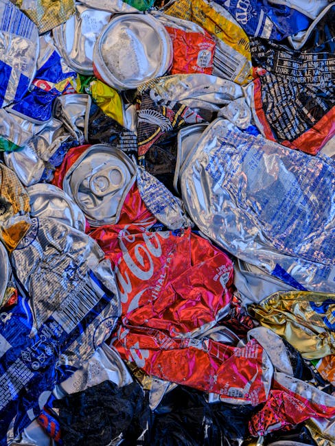 Close-up view of a pile of crushed aluminum cans, including recognizable brands such as Coca-Cola, with some cans partially flattened and others still round, arranged on a surface likely within a household or waste disposal area. The cans show various colors, predominantly red, silver, blue, and black, with some metallic reflections. The scene depicts the recycling or disposal process, which could be part of a home clearance or moving logistics involving waste separation. This image is relevant to house removals and packing procedures handled by [COMPANY_NAME], highlighting the importance of proper waste management during property relocation. The background suggests an indoor environment, possibly a kitchen or storage space, with minimal additional context visible.
