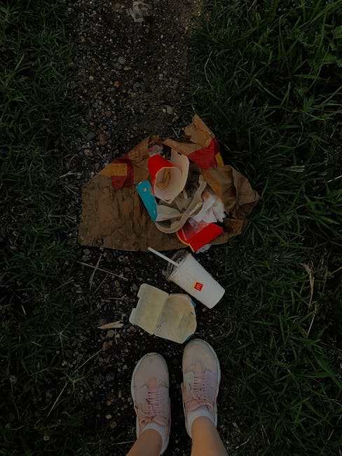 A view of a patch of ground with scattered household waste including crumpled brown paper, a partially crushed red and white cardboard food container, a white plastic bottle with a red logo, and a broken foam food tray. The waste is spread across dirt and gravel, with patches of green grass bordering the area. In the bottom of the image, a person's feet wearing light pink sneakers are visible, standing on the ground near the debris. This scene illustrates improper disposal of household rubbish, which may relate to waste management concerns addressed in house removals and relocation services, such as those offered by Man with Van Shacklewell. Proper refuse handling is an important aspect of moving and packing processes during home relocations.