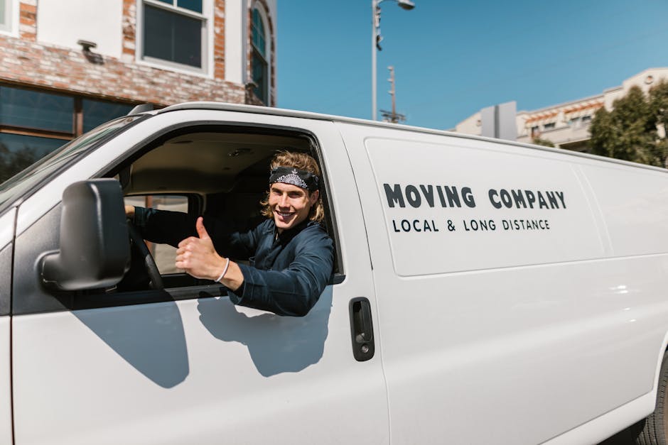 A young male mover with long hair, wearing a black bandana and a dark blue uniform, is sitting in the driverâ€™s seat of a white moving van parked outside a residential building. He is smiling and giv