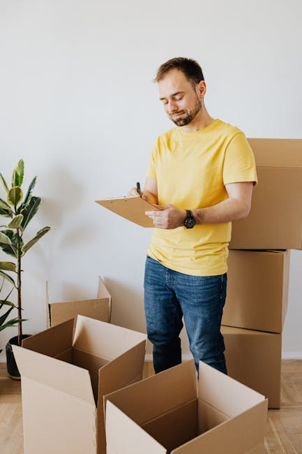 A young man wearing a black bandana, navy blue jumpsuit, and holding a pen is standing inside a room near a cardboard box filled with packing materials. The box contains various items, possibly househ