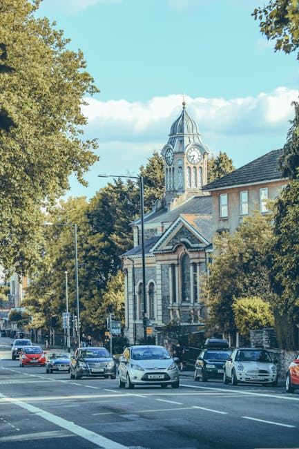 A view of a residential street during daytime with a large, historic clock tower featuring a pointed spire and arched windows in the background. In the foreground, several cars are parked along the curb, and a few are driving down the road. The street is lined with mature trees with green foliage, casting shadows on the pavement. Part of a building with a dark roof and light-colored walls is visible on the right side of the image. The scene suggests a typical urban environment, possibly near a community or town centre, with clear weather and a blue sky with some white clouds. This setting is relevant to house relocations, as it depicts a typical street where furniture loading or moving preparations might occur, and is associated with professional removals services such as those provided by Man with Van Shacklewell.
