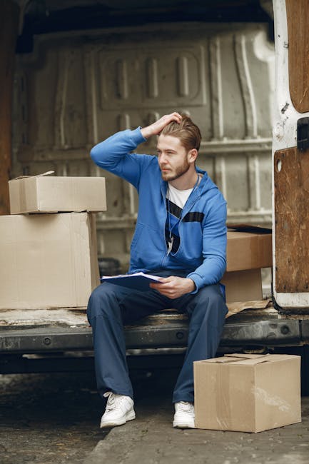 A young man with light brown hair, dressed in a blue zip-up hoodie, white t-shirt, and dark trousers, is sitting on the edge of the open back of a white delivery van. He has a thoughtful expression, with his right hand resting on his head, and is holding a clipboard or notepad in his left hand. Around him, there are several cardboard boxes of various sizes, some closed and others open or partially wrapped. The boxes are placed inside the van and on the pavement beneath him, suggesting an ongoing home relocation or furniture transport process. The van's interior, visible behind the man, shows a grey, textured lining, and the environment appears to be outdoors in a residential or urban setting. This scene depicts the loading or unloading phase of packing and moving, with visible equipment such as boxes and the van used for furniture transport and home removals, consistent with services offered by Man with Van Shacklewell.