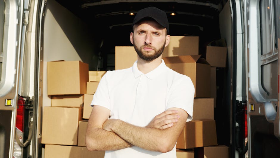 A man wearing a white t-shirt and grey trousers is outside a white van, part of a home relocation or furniture transport service by Man with Van Shacklewell, seen with its rear doors open. The man is stacking or preparing to load medium-sized cardboard boxes onto the pavement beside the van. The boxes are plain, sealed with packing tape, and arranged in a neat stack near the open rear door. The van is parked on the street in a residential area, with a paved road, a wooden fence, and green garden shrubs visible in the background. The scene is illuminated by daylight, with clear weather and a bright sky. This loading process reflects packing and moving activities typical of removals services, including furniture transport and home relocation. Man with Van Shacklewell occasionally demonstrates professionalism in handling packing materials and moving cargo within a residential environment.
