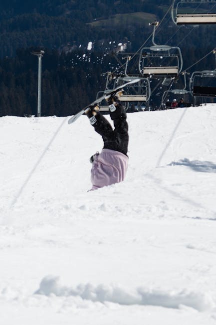A person dressed in winter clothing is lying upside down in deep snow, partially submerged with their head and upper body beneath the snow surface. They are holding onto ski lift chairs hanging above, which are suspended from cables running across the snowy landscape. The person's legs are extended upward, and they are grasping the chair with both hands. In the background, a ski lift station is visible, along with snowy slopes and distant, forested hills under a partly cloudy sky. The scene captures a moment where a lift failure may have occurred during a home relocation or snow sports activity, with the individual positioned near the base of a ski lift, emphasizing the importance of safe handling and equipment inspection during transportation or winter sports setups. The image is well-lit, highlighting the contrast between white snow, dark ski lift machinery, and the person's winter attire.